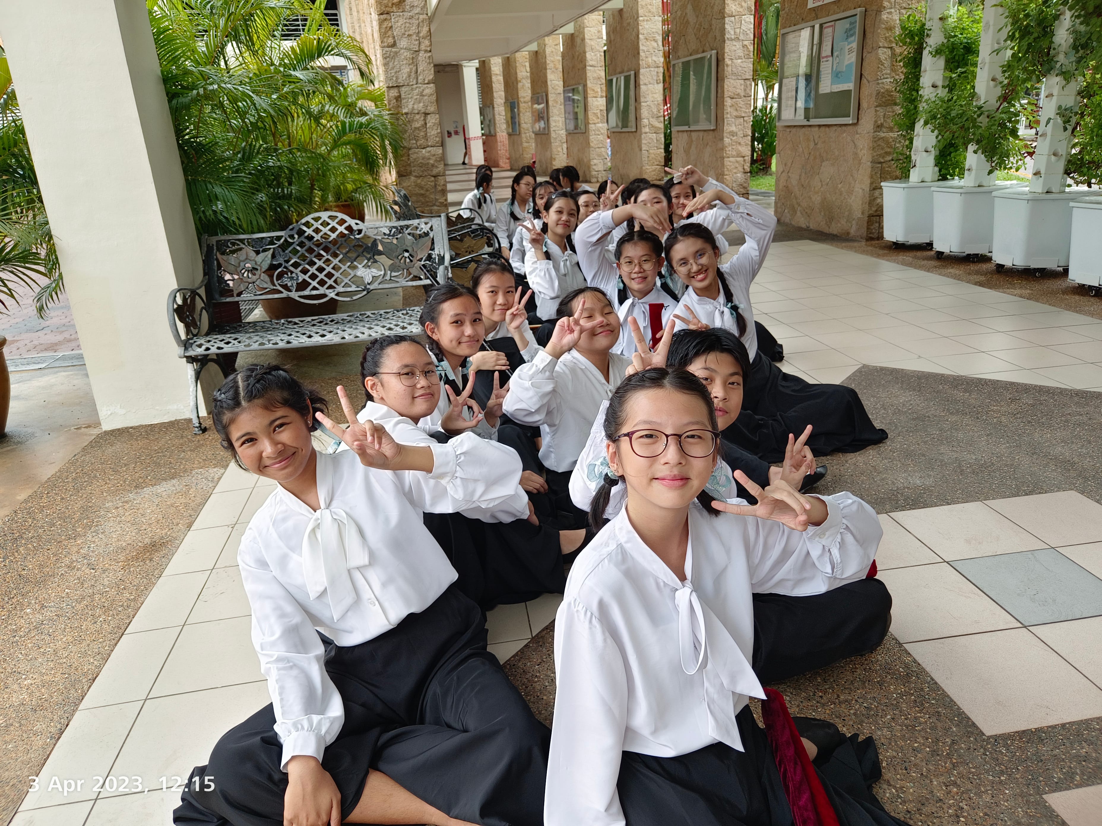 Students in white blouses and dark skirts sit in a line on a tiled walkway, flashing peace signs.